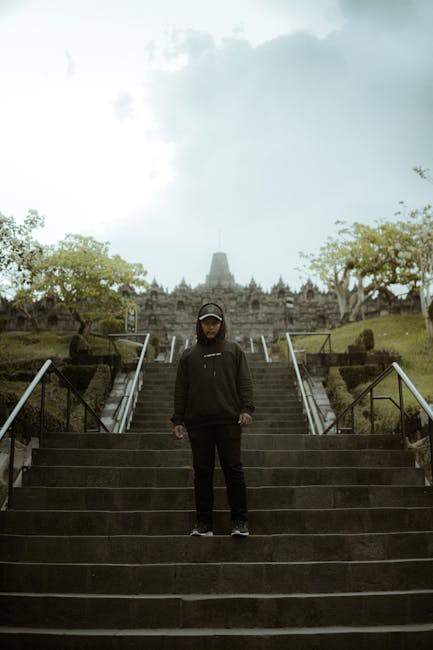 A man in casual attire stands confidently on the steps of a historical site under a cloudy sky.